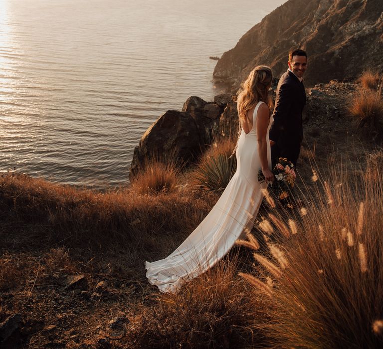 Bride & groom walk along the cliffside on their wedding day as the sun sets across the ocean