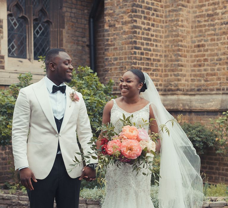 Bride & groom stand outdoors whilst holding hands as bride holds brightly coloured floral bouquet