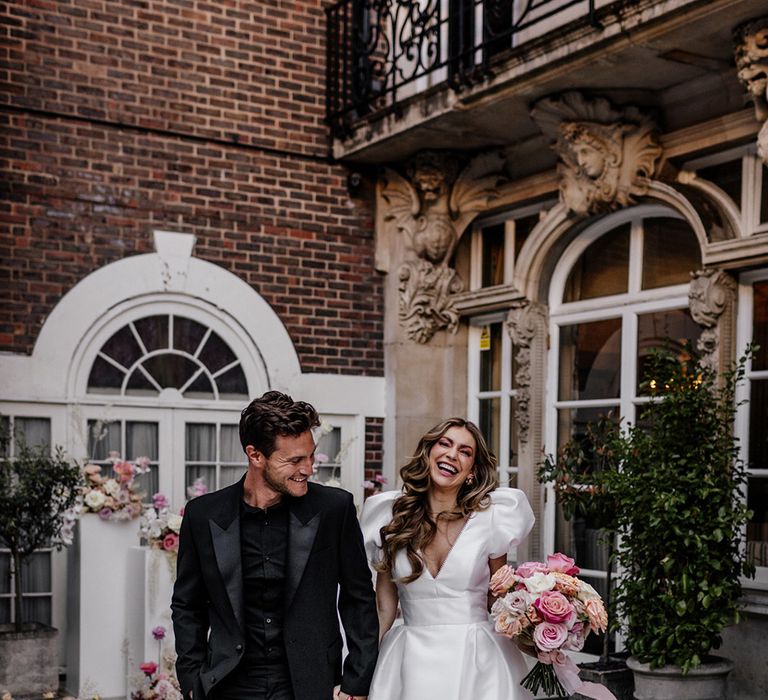 Happy bride and groom in an all black suit, with black shirt and loafers and Bride in a Love in Lace bridal gown with puff sleeves, plunging neckline and open front skirt holding hands 