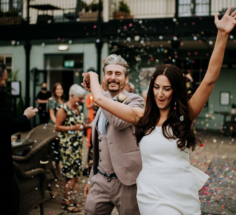 Bride in white strapless Rebecca Vallance Dress throws arm in the air and holds hands with groom in brown Moss Bros suit and blue tie as they walk through multicoloured confetti at the Hotel du Vin after Harrogate wedding