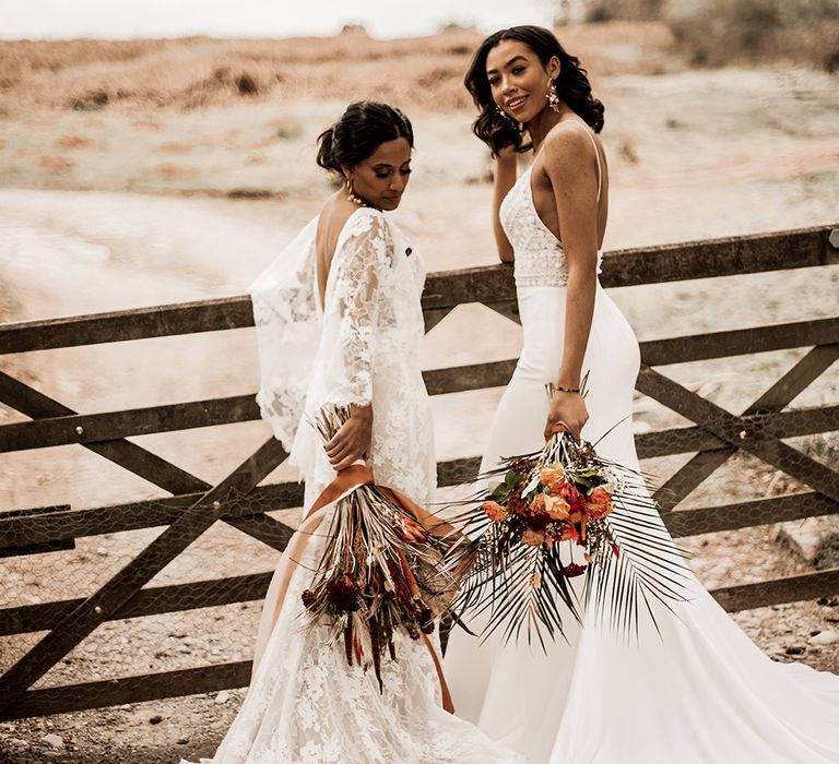 Two bride in, lace and fitted wedding dress holding orange bouquets standing at a field gate 