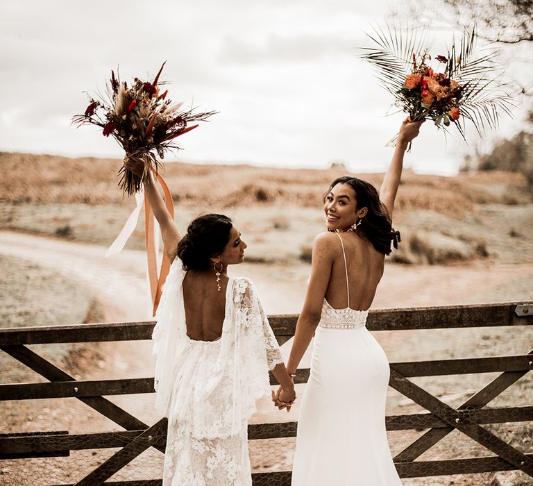 Two brides in a lace wedding dress and fitted wedding dress holding hands and their red bouquets in the air in front of a wooden gate at their farm wedding venue 