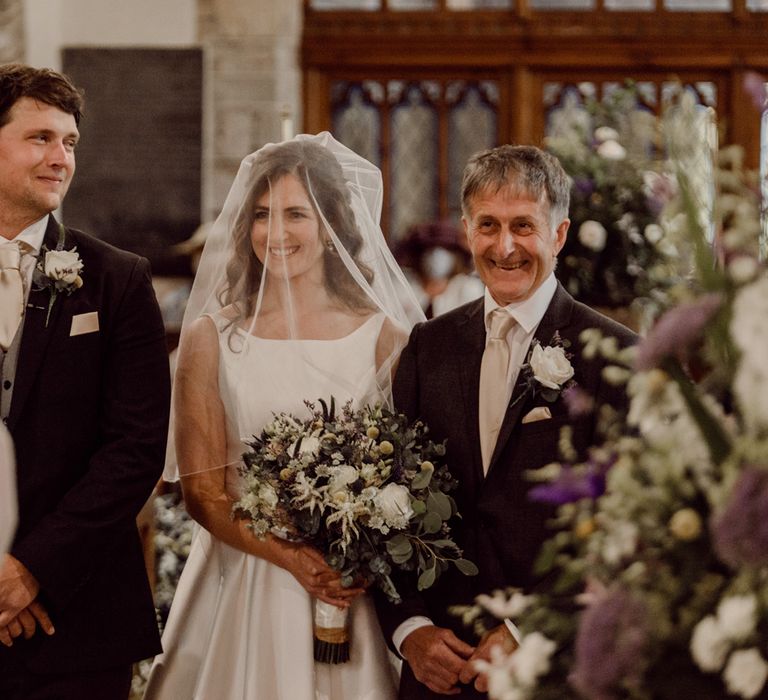 Groom in dark brown suit smiles at bride in Elbeth Gillis wedding gown and father of the bride at the altar of church wedding ceremony