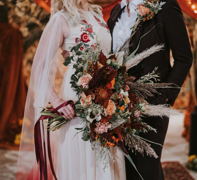 Bride holding a massive bouquet of red and peach roses, carnations, thistles, wild grasses and foliage