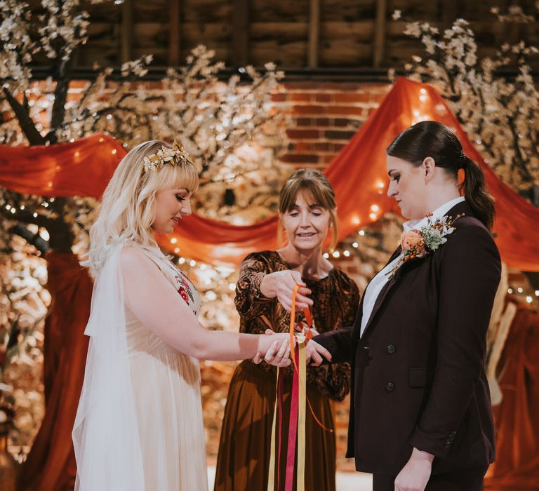 Two brides having a ribbon ceremony with orange ribbons. One bride is wearing a floral embroidered dress with orange-tipped veil and the other is wearing a dark plum coloured suit