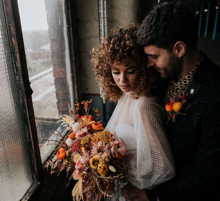 Groom in a patterned shirt embracing his bride in a see through top and bell bottoms holding an autumnal bouquet 