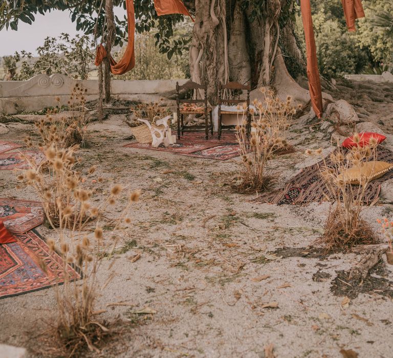 Wedding ceremony setup under secular ficus tree, complete with Moroccan style rugs for sitting, orange fabrics hanging from the tree, and two chairs for bride & groom 