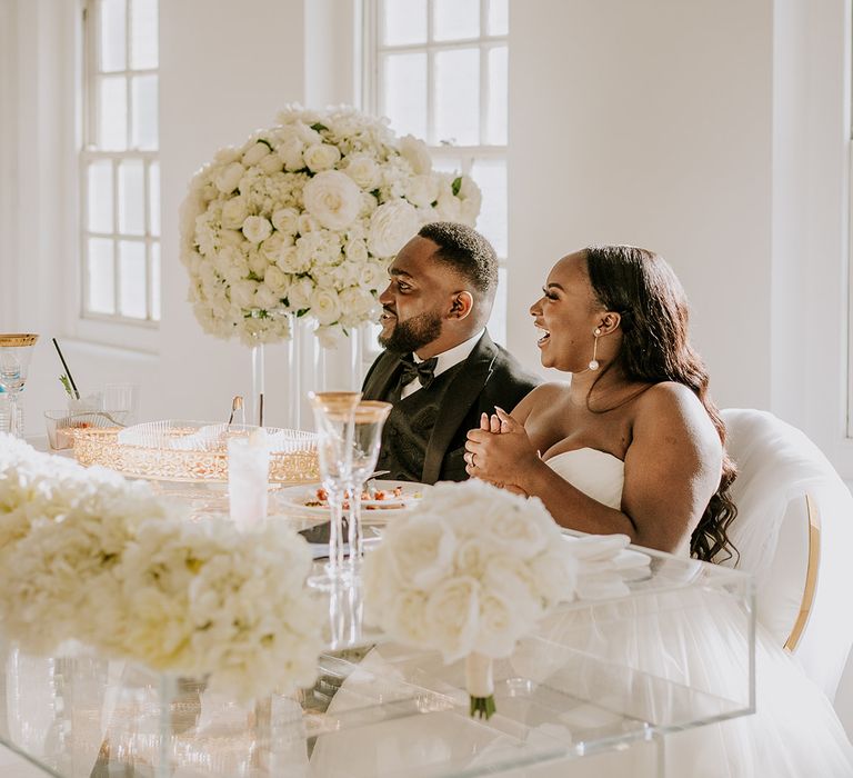 Bride & groom sit together during reception surrounded by white roses in large floral bouquets 