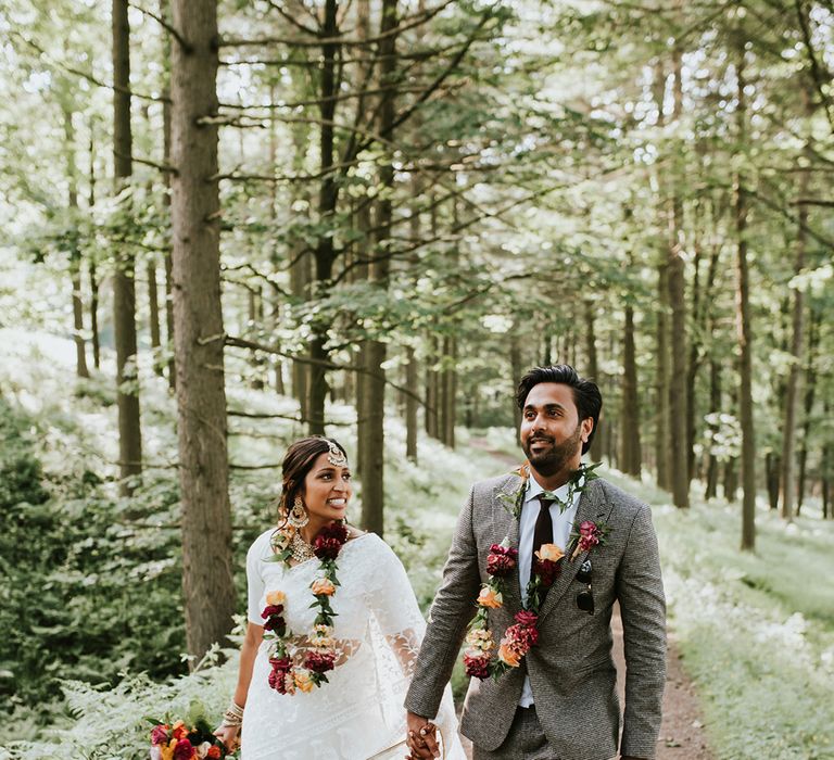 An Indian British bride and groom walk through woodland holding hands. He wears a grey suit and she wears a white sari.