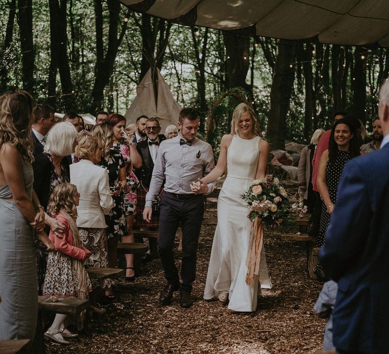 Bride & groom hold hands as they walk through woodland as wedding guests look on and smile