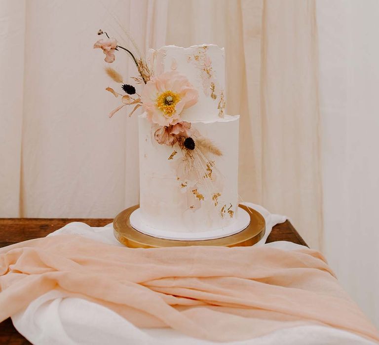 Two tier buttercream wedding cake decorated with gold leaf and dried and fresh flowers on a gold cake stand 