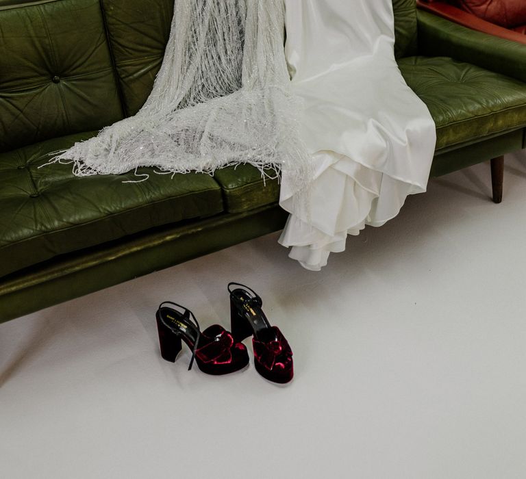 Halfpenny London dress and bridal cape laid on green leather sofa next to burgundy velvet block heels for wedding at Loft Studios London