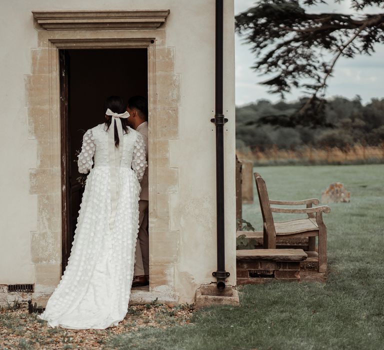 Bride in long mesh sleeve spotted Vagabond wedding dress and bridal hair bow stands in church doorway for Wasing park wedding