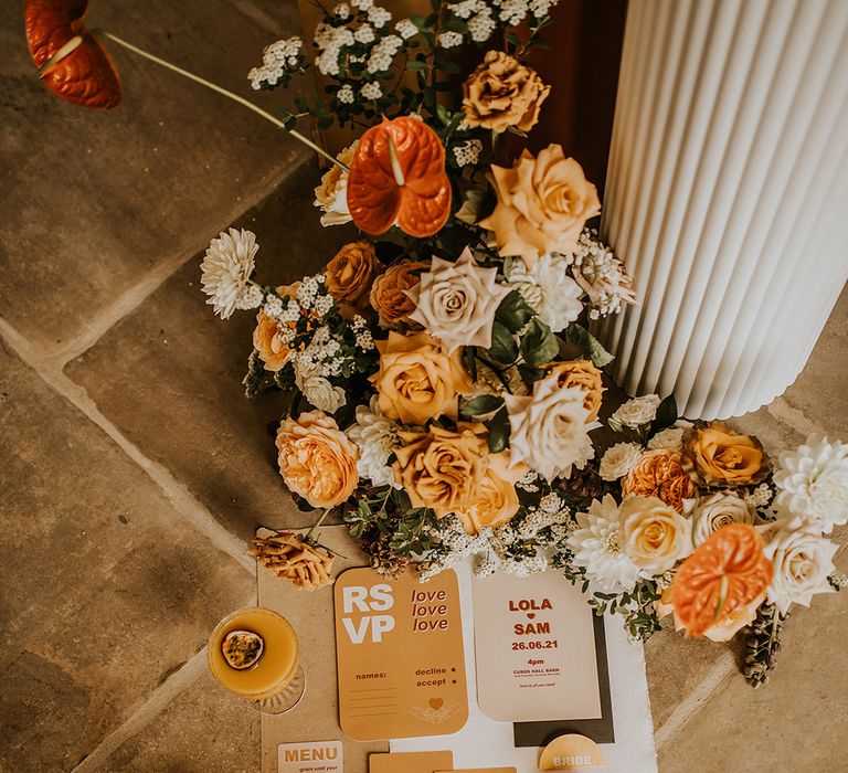 Flat lay of contemporary mustard yellow and burnt orange wedding stationery suite surrounded by anthurium and roses floral arrangement 