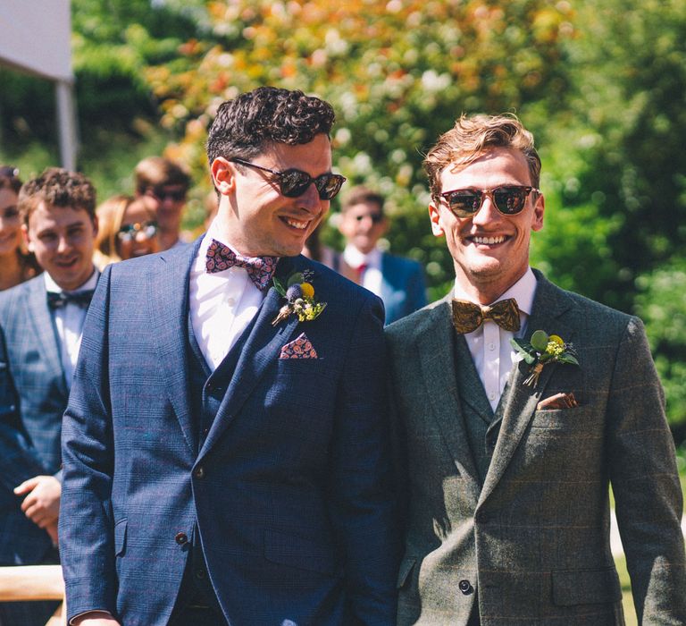 Grooms walk down the aisle wearing modern suits complete with floral buttonholes and sunglasses  | Story + Colour