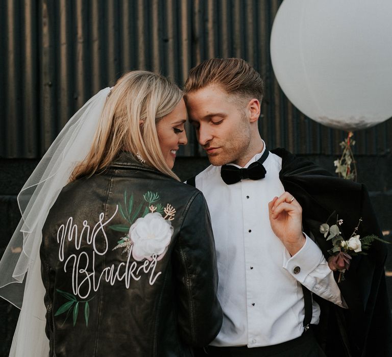 Groom in a tuxedo holding a giant balloon embracing his bride in a hand-painted personalised leather jacket with name and floral design 