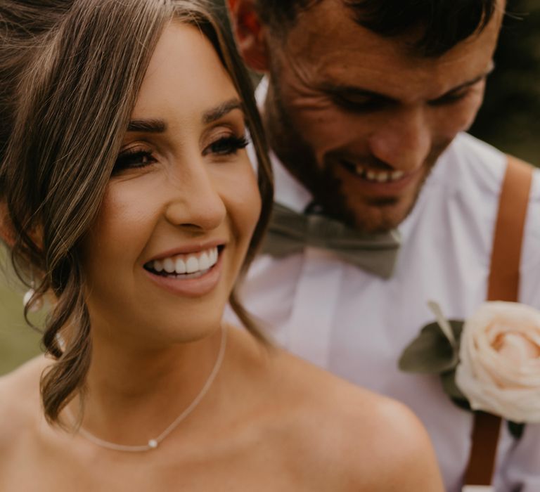 Bride laughs as her groom stands behind her wearing white shirt and white floral buttonhole | Mark Bamforth Photography