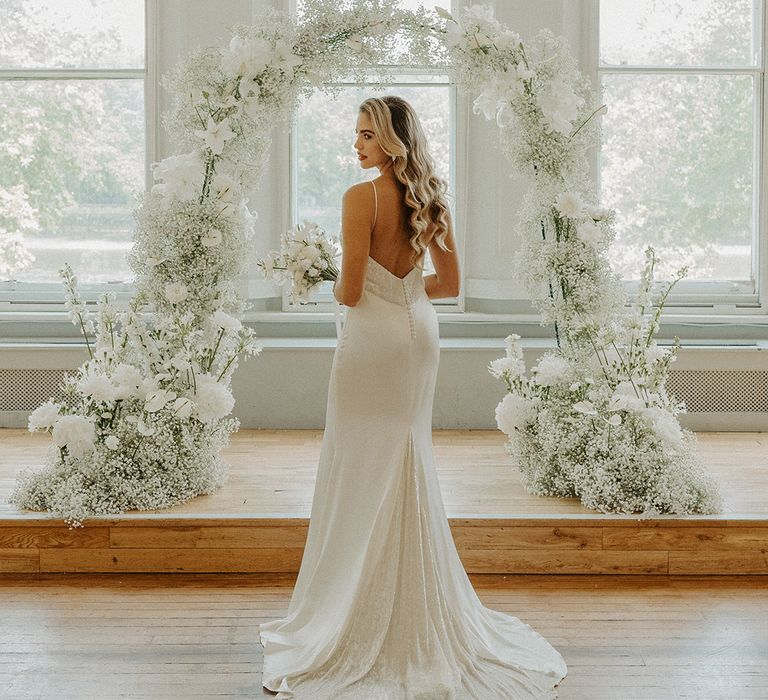 Bride in a slip wedding dress with embellished bodice and train standing in front of a white flower and gypsophila flower arch 