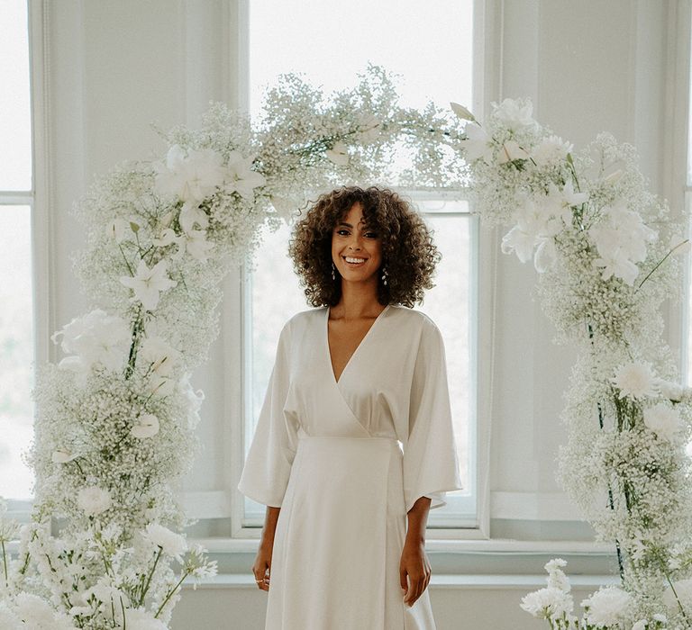 Black bride with naturally curly hair in a wrap satin wedding dress by Consellation Ame standing in front of a white flower and gypsophila flower arch 