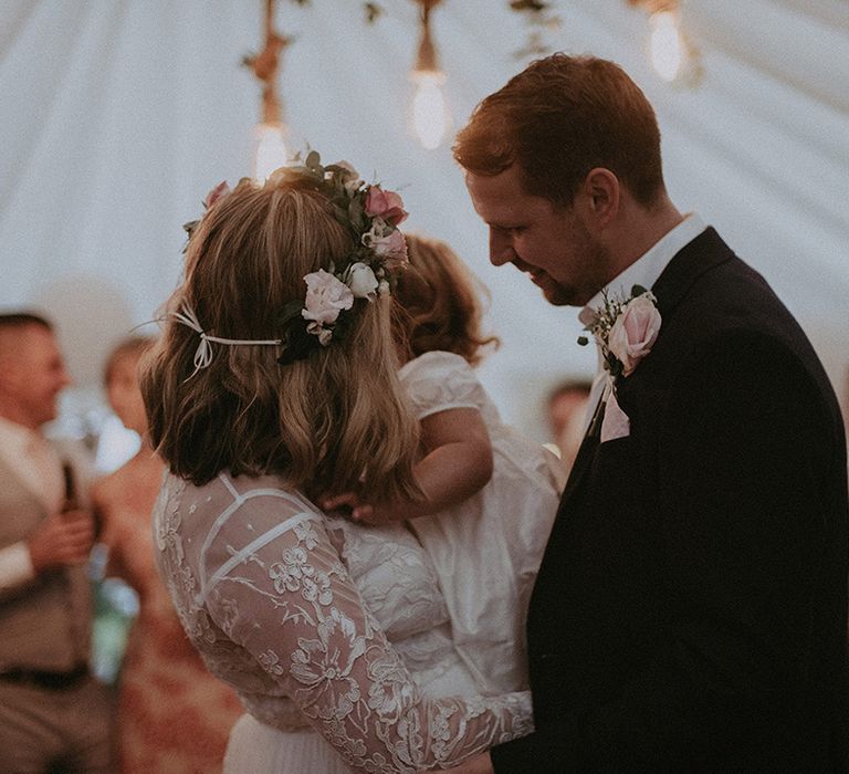 Bride with lace long sleeves holding her daughter on the dance floor with her husband in a navy blue blazer at their intimate home wedding 