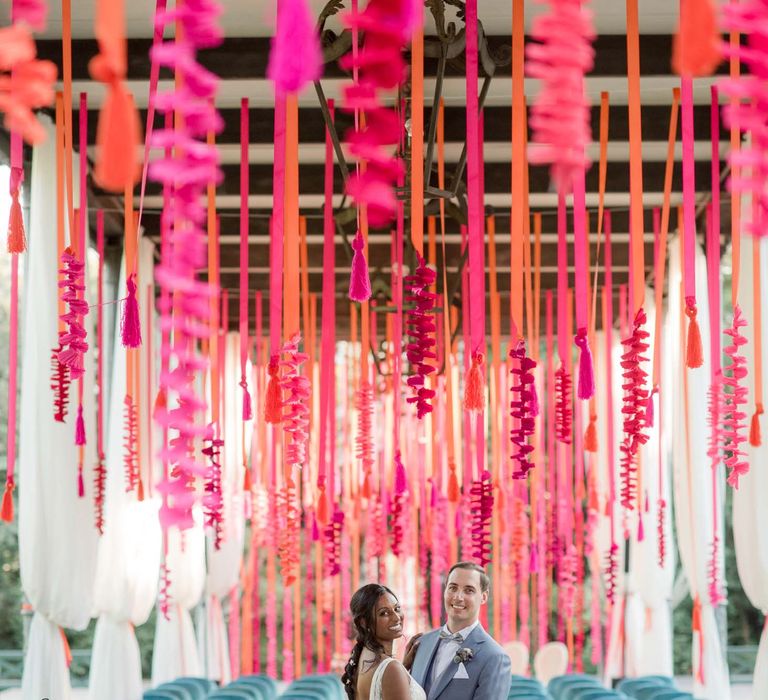 Bride & groom stand underneath colourful hangings surrounded by emerald blue velvet chairs