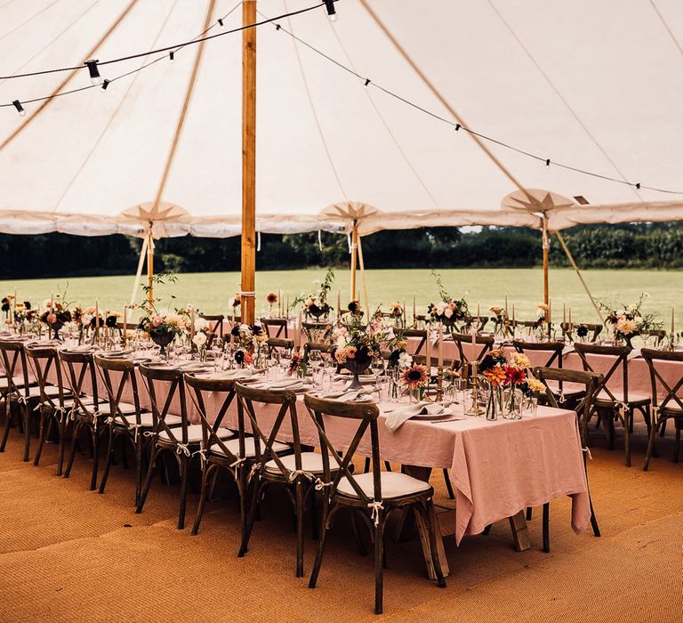 Two long wooden tables with dusky pink tablecloths, candles and assorted florals for marquee garden wedding reception in Cornwall