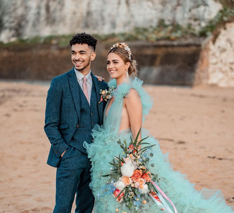 Bride and groom portrait at their coastal elopement with bride in a mint green wedding dress holding a pastel wedding bouquet tied with ribbon and groom in a blue wool suit 