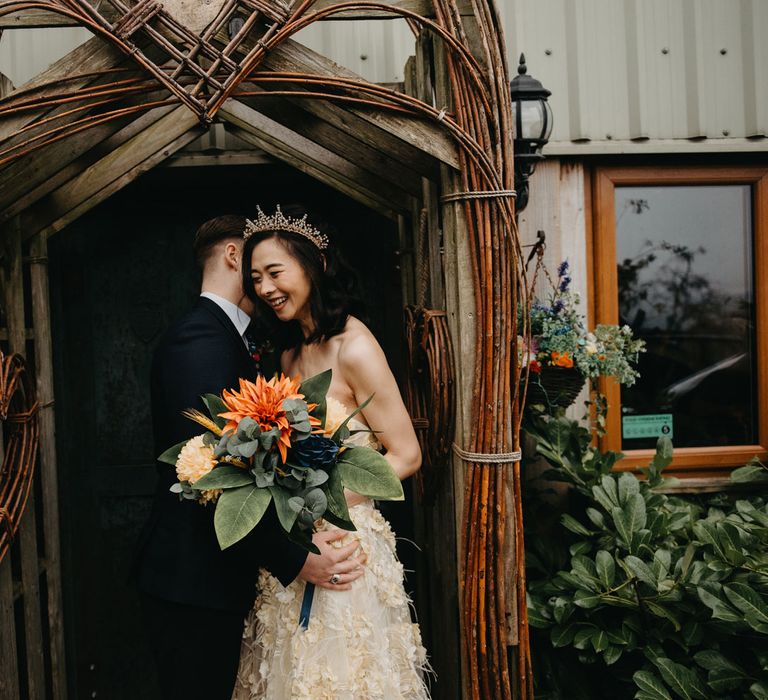 Laughing bride in strapless feathered wedding dress and tropical wedding bouquet stands with groom in dark suit in doorway for farm wedding in Lancashire