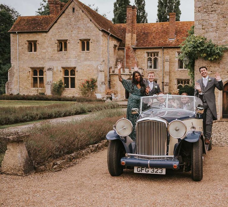 Groom, groomsmen and woman in sequin green dress wave from vintage car parked in the grounds of Notley Abbey in Buckinghamshire 