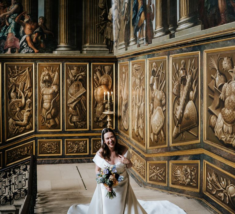 Bride stands within beautifully gilded room on her wedding day whilst laughing and holding floral bouquet