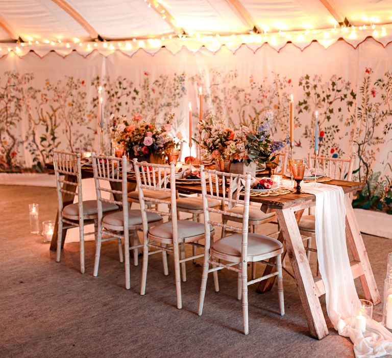 Interior of modern marquee tent for wedding reception with wooden table and chairs, bright florals and pastel candles for boho wedding