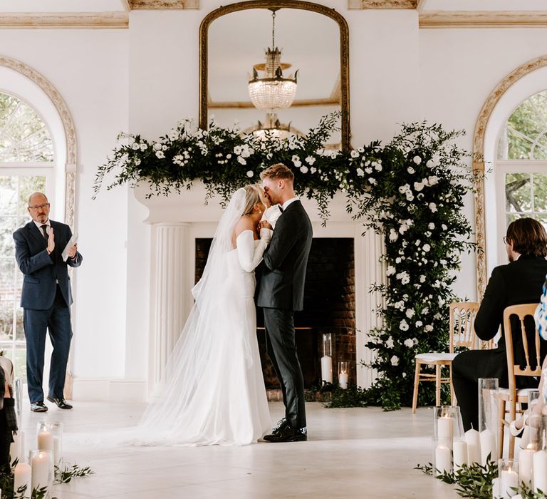 Wedding ceremony at Northbrook Park with Bride in a fitted wedding dress and groom in a tuxedo kissing in front of the fireplace decorated with green foliage and white flowers