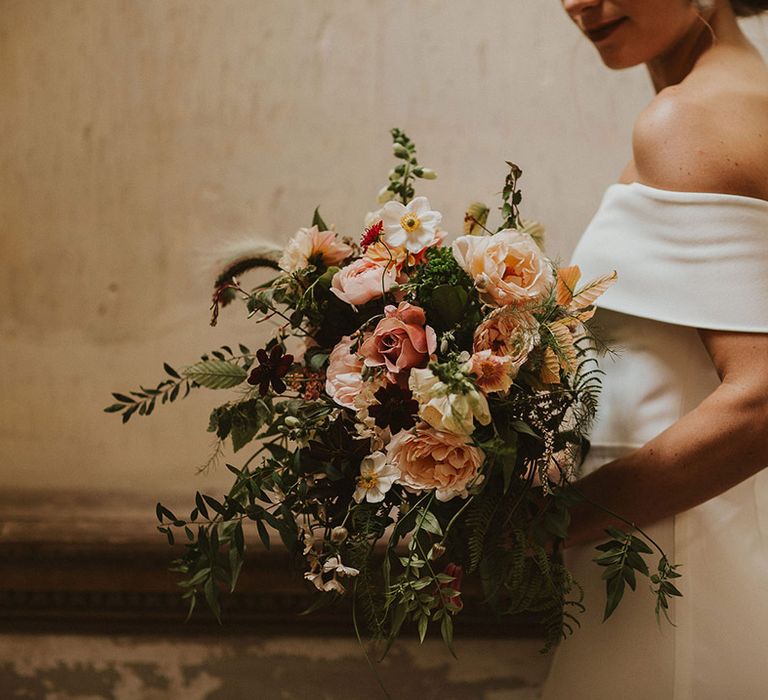 Bride wears off the shoulder wedding gown whilst holding peach and pale pink floral bouquet