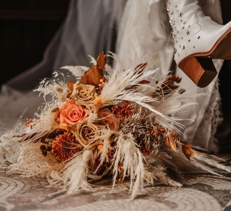 Bride in white cowboy boots sits next to mixed orange and white bridal bouquet with roses and dried florals