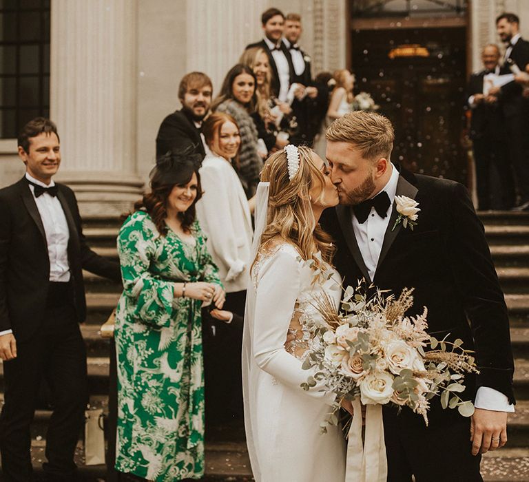 Bride in a long sleeve wedding dress holding a romantic wedding bouquet kissing her groom in a tuxedo at the bottom of the Old Marylebone Town Hall steps 