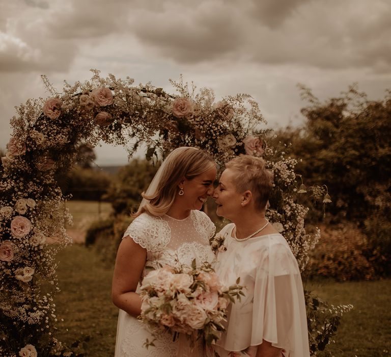 Bride in a lace wedding dress with her mum in front of a pink rose and white gypsophila flower arch 