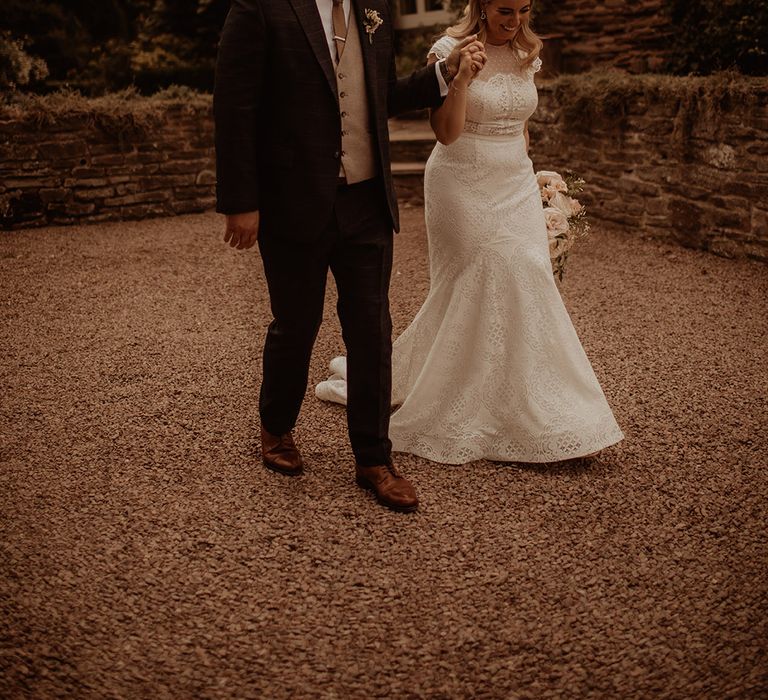 Groom in a three-piece wedding suit holding hands with his bride in a lace fishtail wedding dress with cap sleeves