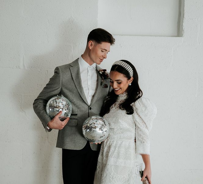 Bride wears short wedding gown as she stands with groom in grey blazer who both carry disco ball decoration