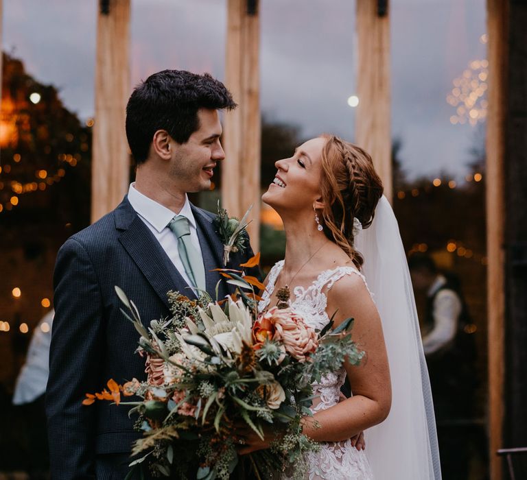 Bride with braided updo and veil holding large mixed bridal bouquet smiles at groom in dark suit and light grey tie outside barn