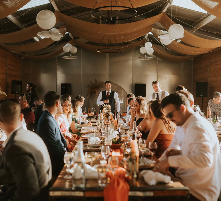 Wedding guests sit at long wooden tables with orange rustic wedding table decor during barn wedding reception at Hope Farm in Dorset