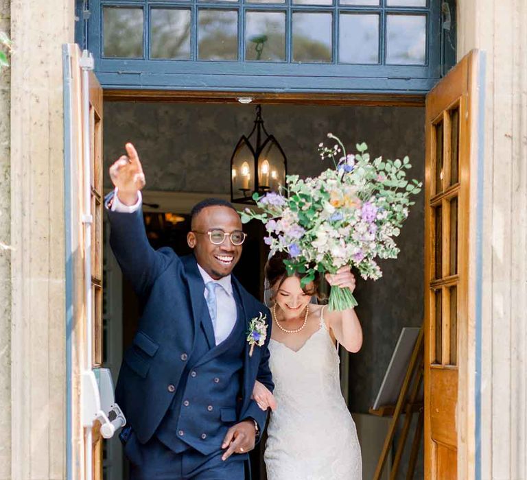 Bride lifts pastel bouquet in the air as she walks out the door with her groom