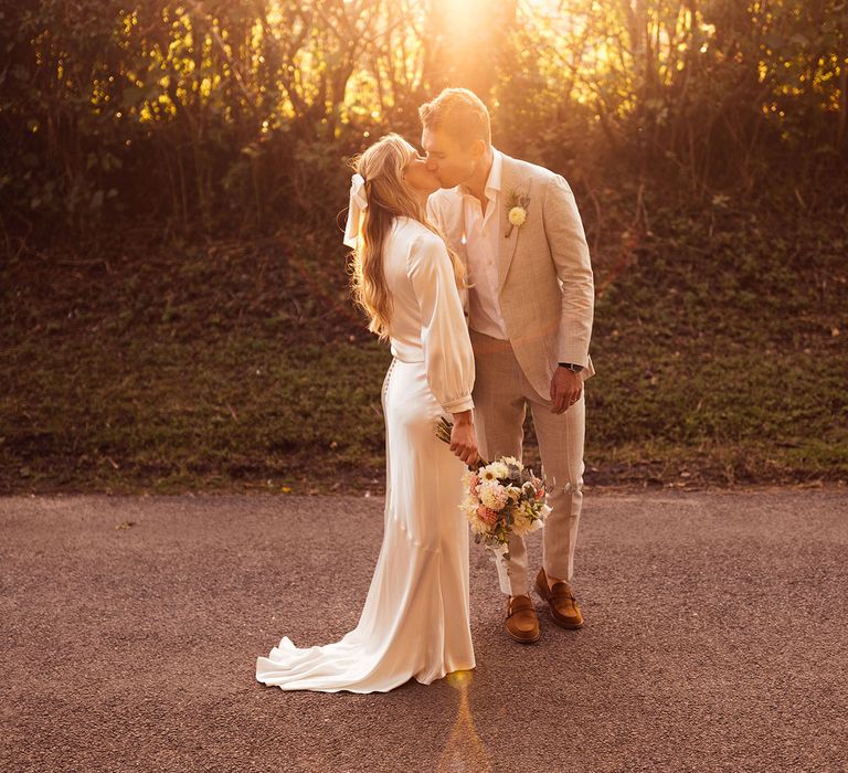 Bride in satin dress with button detailing and train holding pink and yellow bridal bouquet kisses groom in grey suit and floral buttonhole on country lane during golden hour