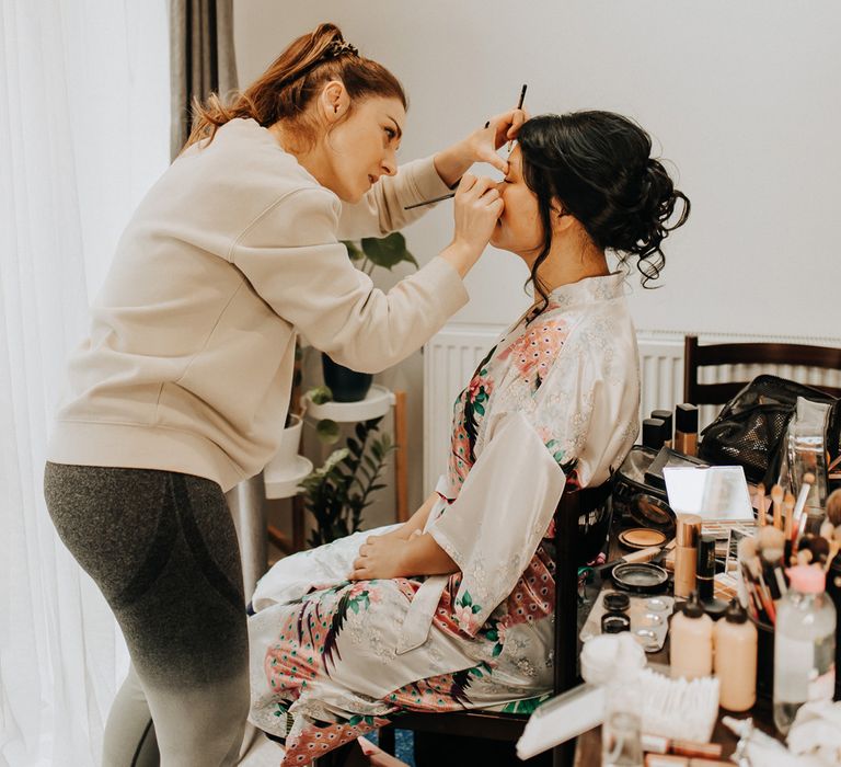 Bride in patterned silk kimono dressing gown sits having her makeup done before wedding