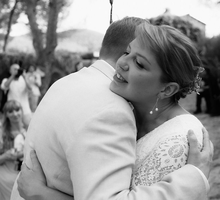 Bride in lace wedding jumpsuit with diamanté belt and dainty pearl earrings hugs wedding guest