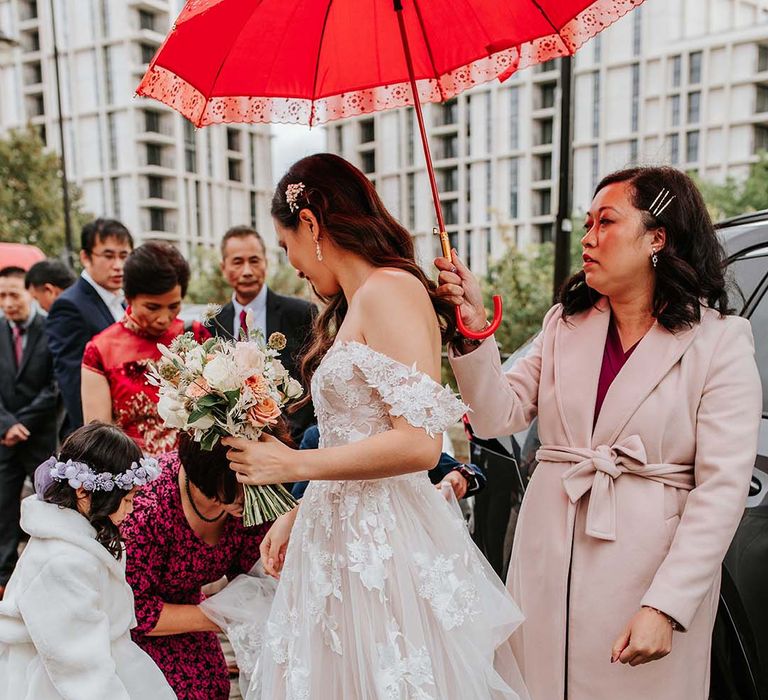 Bride in tulle sweetheart wedding dress with lace flowers and off the shoulder detail stands under a red umbrella with lace trim 