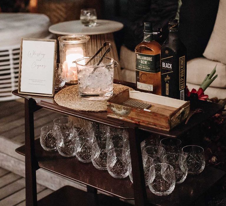 Whiskey and cigar station on bar cart with sign in gold frame, ice bucket and wooden case of cigars