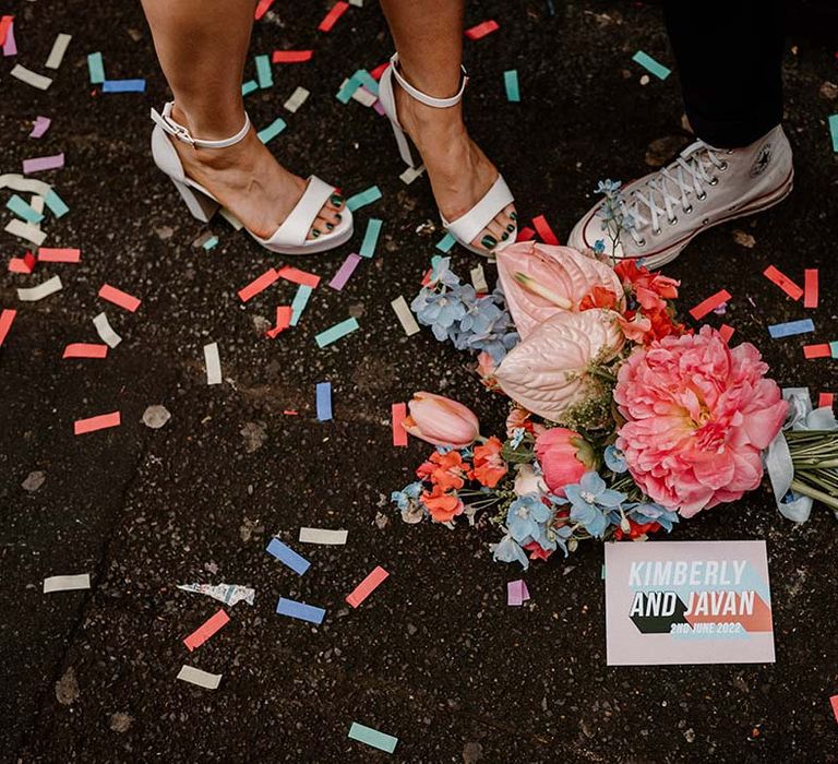 Bride in white platform shoes and groom in Converse high tops standing next to a pink and blue wedding bouquet 