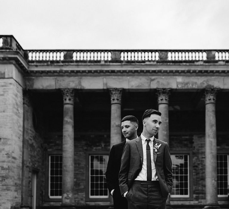 Grooms pose together and stand in front of the other in front of venue 