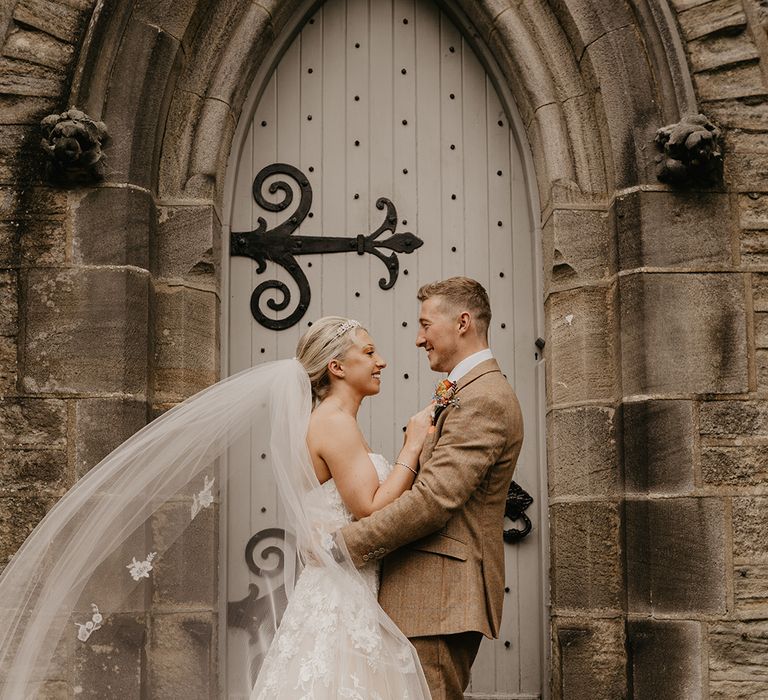 blonde bride in strapless sweetheart lace wedding dress and embroidered veil with groom in beige suit in front of door