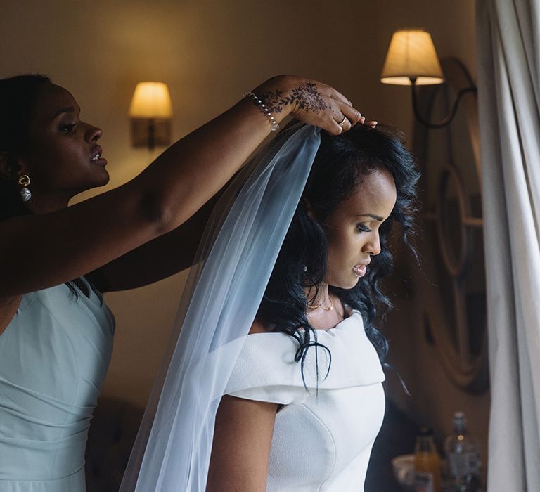 Bridesmaid in mint green dress and pearl earrings helps put on the bride's veil 